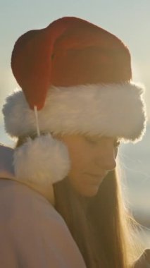 Dikey video. Noel sabahı deniz kenarında Noel Baba şapkalı genç bir kadın zencefilli kurabiye ve çayın tadını çıkarıyor. Arkasında parlak güneş.