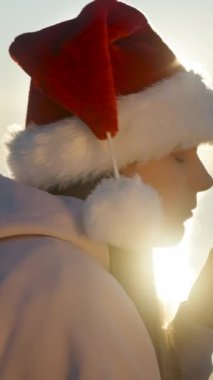 Dikey video. Noel Baba şapkalı genç bir kadın Noel 'i deniz kenarında sıcak çay ve zencefilli şekerle kutluyor..