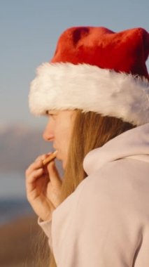 Dikey video. Genç bir kadın gün doğumunda deniz kenarında oturur, Noel kurabiyesi ve çayın tadını çıkarır, arkasında dağlar vardır..