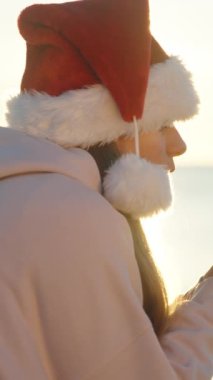 Dikey video. Deniz kenarında serin bir Noel sabahı, kırmızı Noel Baba şapkalı genç bir kadın sıcak çay içiyor. Kadehten buhar yükseliyor..