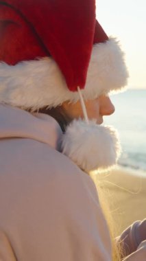 Dikey video. Deniz kenarında serin ama güneşli bir Noel sabahı. Noel Baba şapkalı genç bir kadın ısınıyor, elinde bir fincan sıcak çay tutuyor..