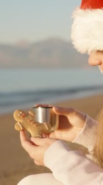 Dikey video. Noel Baba şapkalı genç bir kadın, zencefilli kurabiyelere erkek şeklinde bakar ve elinde bir kupa çay ile şafak vakti deniz kenarında oturur..