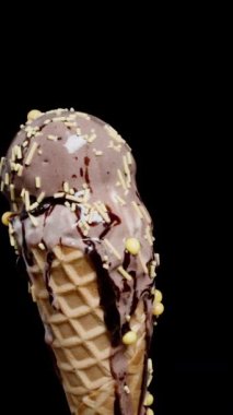 Dikey video. Çikolatalı dondurmalar waffle külahında şekerli şekerlemeler dönüyor ve eriyor, siyah bir arka plana karşı..