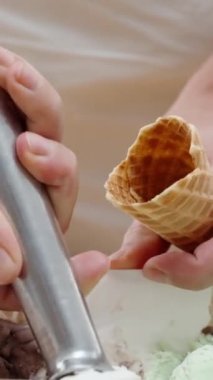 Dikey video. Kadının elleri özel bir kaşıkla waffle külahına dondurma topları atıyor..