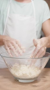 Bir kadın modern bir mutfağın arka planında kiraz galonu için cam bir kasede hamur yoğuruyor. Beyaz bir önlük giyiyor. Dikey video.