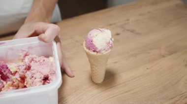 Kadın özel bir kaşıkla dondurmayı kutudan topluyor ve yanında da pembe dondurma külahlı waffle külahı var..
