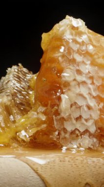 Dikey video. Ahşap bir tahtanın üzerindeki bal peteklerinde mühürlü bal damlıyor, yakın plan, siyah arka plan üzerinde..