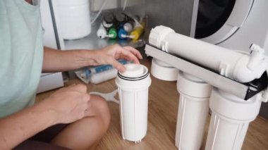 Genç bir kadın mutfakta yerde oturuyor ve su filtrelerini değiştiriyor. Beyaz konutlu ters osmoz sistemi..