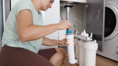 Genç bir kadın bağımsız olarak su filtrelerini değiştiriyor. Mutfak zemininde otururken beyaz bir kapaktan mavi bir zar filtresi çıkarıyor..