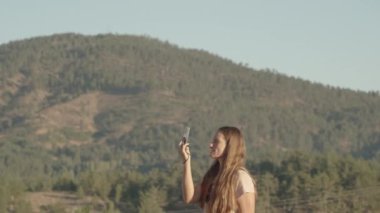 Uzun saçlı genç bir kadın cep telefonunda ağaçlarla güzel dağ manzaraları yakalıyor..