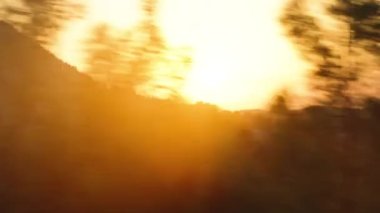 Yürüyen bir arabadan engebeli dağ yolları boyunca bir manzara, önemli bir sarsıntı, ve gün batımı ağaçların arasından gözetliyor..