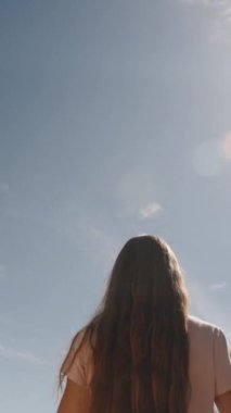 Dikey video. Gezgin genç kadın, antik harabelerin ve kayaların arasında saçlarını güneşin ve yüksek dağların arkasında, eliyle topluyor..