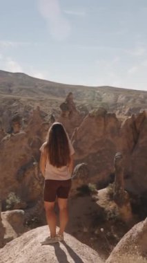 Dikey video. Genç bir kadın yüksek uçurumların arasındaki büyük bir kayanın üzerinde duruyor ve ben kayanın arkasından bakıyorum. Hayal gücü vadisi..
