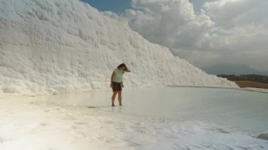 Pamukkale 'nin beyaz travertinelerinde bir kadın suyun üzerinde yürüyor. Arka planda, beyaz kalsit yataklarından yapılmış büyük bir duvar var..