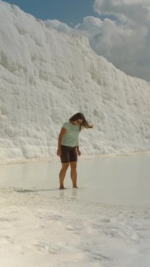 Dikey video. Pamukkale 'nin beyaz travertinelerinde bir kadın suyun üzerinde yürüyor. Arka planda, beyaz kalsit yataklarından yapılmış büyük bir duvar var..