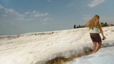 Pamukkale 'nin beyaz geçitlerinde beyaz uçurumun kenarındaki su kanalı boyunca yürüyen bir kadın. Panorama.