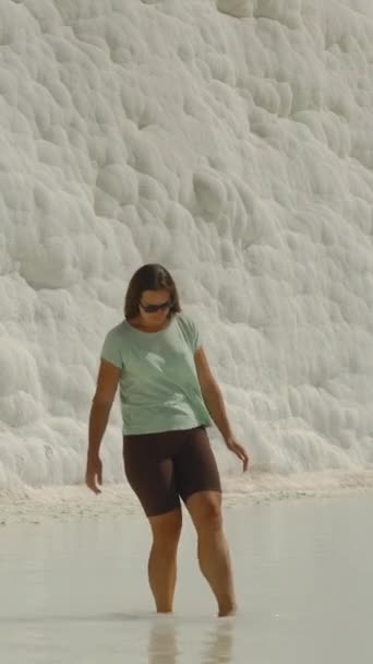 垂直录像 一个年轻的女人在帕穆克卡莱的白色长裙内的泉水中浸泡着她的脚 在背景中 有一堵白色的墙 上面有方解石矿床 展示着古代的地层 — 图库视频影像