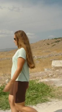 Dikey video. Genç bir kadın, güneşli bir günde, dağların ve çalıların arkasında, antik taş kiremitlerden oluşan bir yamaçta yürüyor..