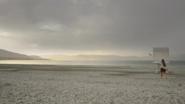 Hava bulutlu, güneş dağın arkasında batmak üzere ama yaklaşan yağmura rağmen genç bir kadın göle doğru yürüyor..