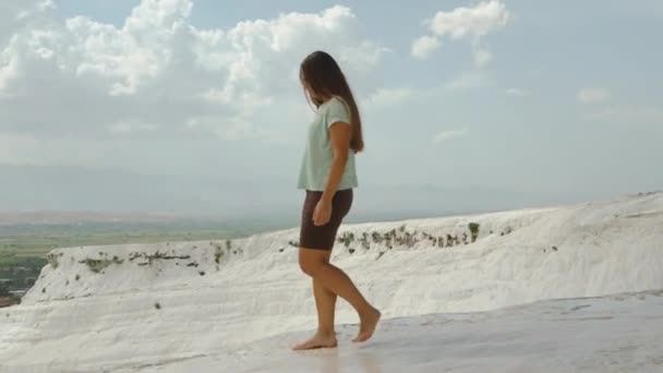 若い女性が白いカルシウムの堆積崖の端に裸足を歩いています 山の中では高い パムッカレのトラバーチン 空の壮大な雲 — ストック動画