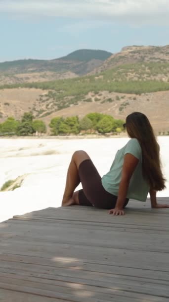 垂直录像 一个年轻的女人坐在白山顶上的一座木桥上 Pamukkale的Travertines 她欣赏平原和群山 相机以圆形的运动运动 — 图库视频影像