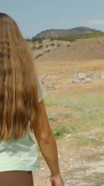 Dikey video. Dağın eteğindeki eski bir şehrin kalıntıları arasında eski taş yolda yürüyen genç bir kadının manzarası..
