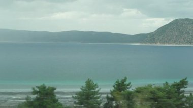 Bir dağ gölünde turkuaz su, çorak kıyılar ve uzak dağlar. Araba penceresinden manzaralı bir yolculuk..