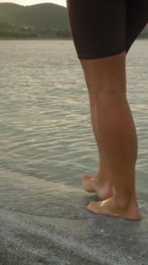 Dikey video. Gün batımının son ışınları Dağ Gölü 'nün sularına düşer. Genç bir kadının çıplak ayakları suda yürürken..