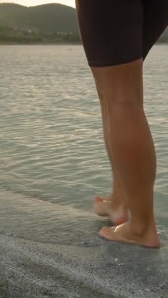 バーティカルビデオ サンセットの最後のレイズは 若い女性の裸足が水の中を歩くように マウンテンレイクの水に落ちます — ストック動画