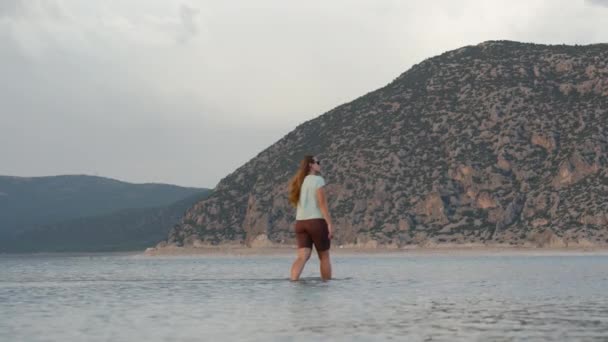 若い女性が山岳湖の浅瀬を通り抜け 天候が荒れ 遠くの山々を背景に歩いている スローモーションで — ストック動画