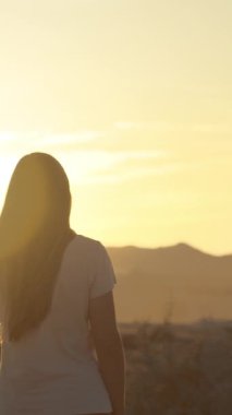 Dikey video. Batan güneşin sıcak ışığı yamaçta yürüyen kadını aydınlatır. Ağır çekimde.