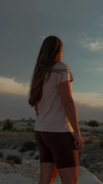 Dikey video. Yalnız genç bir kadın bir dağın tepesinde kanyona tepeden bakıyor ve uzak bir dağın ardında günbatımının tadını çıkarıyor. Dairesel hareket.