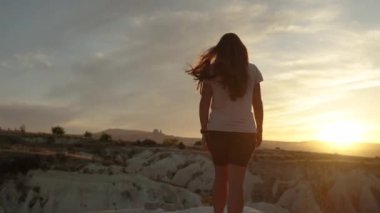 Genç bir kadın gün batımında dağın kenarında yürüyor, altında derin bir kanyon var..