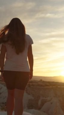 Dikey video. Genç bir kadın gün batımında dağın kenarında yürüyor, altında derin bir kanyon var..