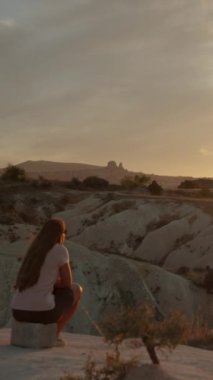 Dikey video. Bir kadın yüksek bir dağdaki uçurumun kenarında oturuyor, günbatımının tadını çıkarıyor. Beyaz kayalıklar.