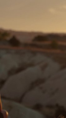 Dikey video. Genç bir kadın beyaz bir uçurumun kenarında oturuyor, gün batımını seyrediyor, odak yavaşça ona yaklaşıyor..