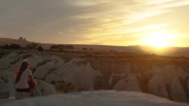 Genç bir kadın bir dağın yamacında oturuyor, günbatımının tadını çıkarıyor, sakinliğini ve yalnızlığını ifade ediyor, sadece rüzgarın hafifçe saçlarını okşamasıyla.