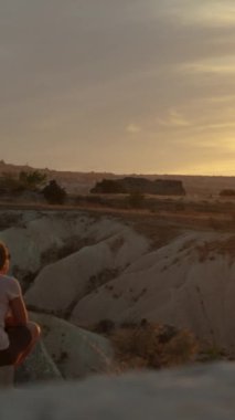 Dikey video. Genç bir kadın bir dağın yamacında oturuyor, günbatımının tadını çıkarıyor, sakinliğini ve yalnızlığını ifade ediyor, sadece rüzgarın hafifçe saçlarını okşamasıyla.