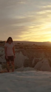 Dikey video. Güneş dağın arkasında batmak üzere ve kadın uçurumun kenarından kanyona doğru yürüyor..