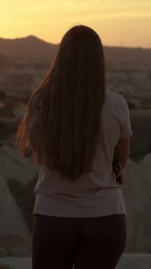 Dikey video. Bir uçurumun kenarında, yalnız bir kadın, dağlarla ve önünde beyaz bir kanyonla duruyor, ve güneş dağın arkasında uzaklardan batıyor..