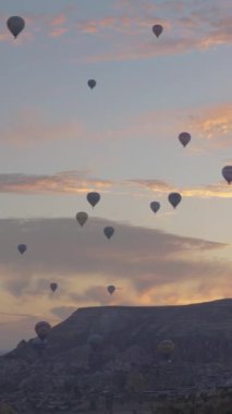 Dikey video. Yüzlerce sıcak hava balonunun insanları havada taşıdığı kanyonda güzel bir gün doğumu. Güneşli altın bulutların ve yükselen uçurumların ardında..