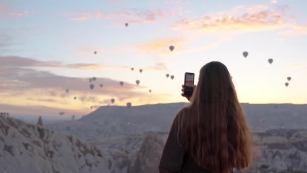 在日出时分 一位女士在手机上捕捉到美丽的景色 数百个热气球在高山上空飞舞 — 图库视频影像