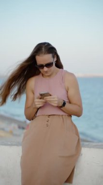 Dikey video. Yavaş çekimde rüzgarda sallanan saçlar. Arka planda deniz olan telefonuna bakan genç bir kadın..