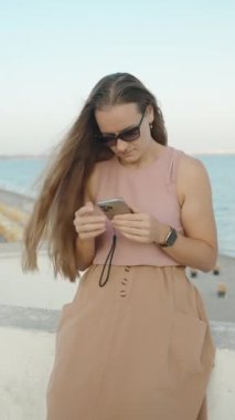 Dikey video. Sahil İskelesinde genç bir kadın, telefonundan bir şeye bakıyor, ağır çekimde..