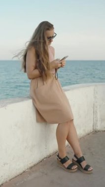 Dikey video. Deniz kenarındaki Beton İskeledeki Çıplak Etekli Genç Bir Kadın Yavaş Çekimde telefonuna bakıyor..