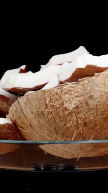 Dikey video. Kahverengi bir hindistan cevizi parçalara ayrılır ve beyaz et cam bir kapta dizilir. İzole edilmiş bir arkaplanda bir döngü içinde dönüyor.