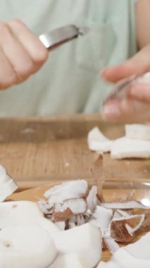 Dikey video. Sebze soyacağı kullanan kadın, kahverengi deriyi hindistan cevizi etinden çıkarıp tabağa yerleştirir. Makro Kaydırıcı.