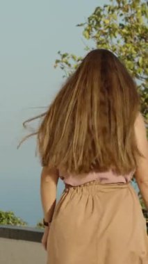 Dikey video. Arka planda deniz ve mavi gökyüzü olan ağaçların arasında yürürken rüzgarda süzülen uzun saçlı genç bir kadının manzarası..