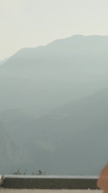 Dikey video. Genç bir kadın uçurumun kenarına yaklaşıyor. Dağların ve aşağıdaki denizin görkemli manzaralarını geziyor ve hayranlık duyuyor. Batan Güneş uzun saçlarını aydınlatıyor..