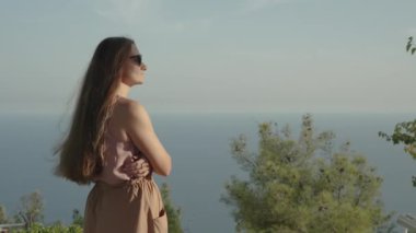 Daire çizen sinematik kamera, dağın tepesinde genç bir kadın yakalıyor, günbatımının ve denizin manzarasının tadını çıkarıyor. Yavaş Çekimde.
